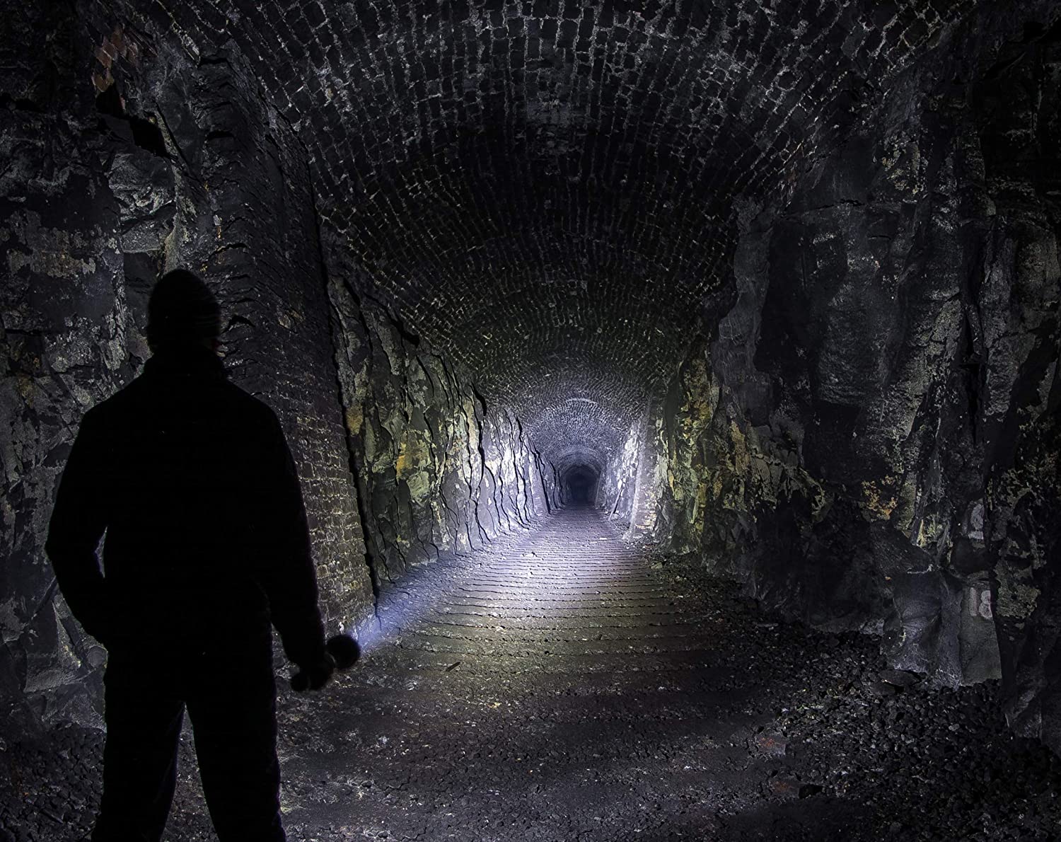 Eine Person mit einer LEDLENSER Ledlenser Lampe X21R steht am Eingang eines dunklen, felsigen Tunnels und beleuchtet die rauen Steinwände und die Ziegeldecke, während sich der Tunnel in die Ferne erstreckt.