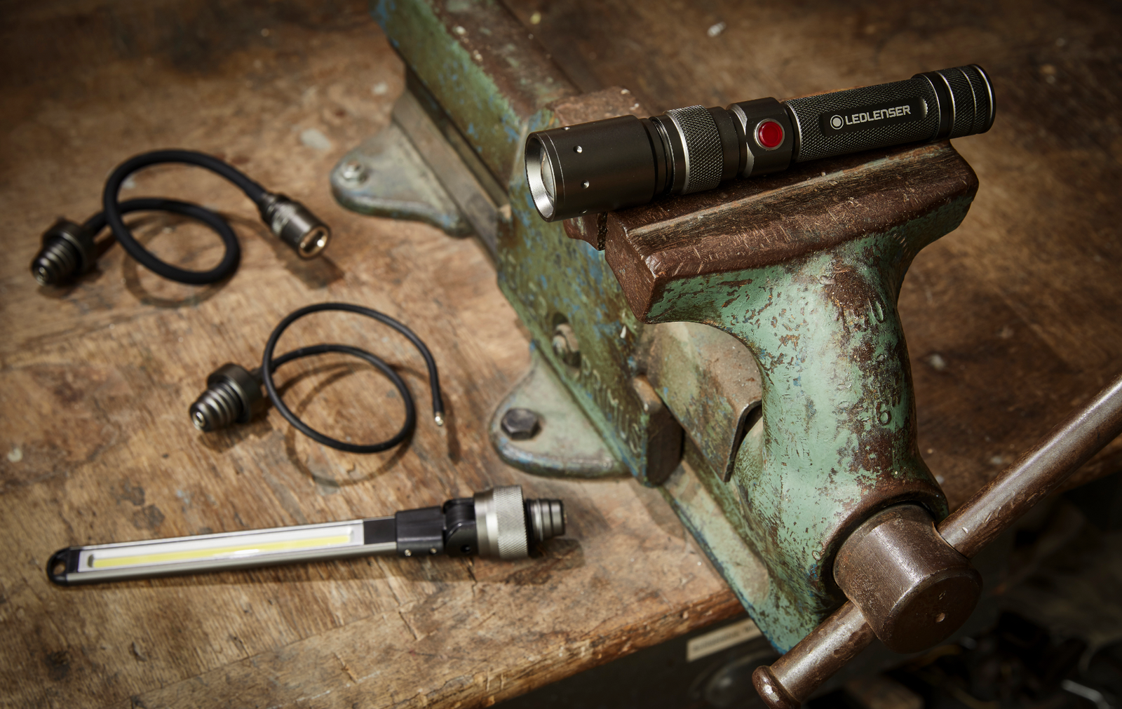 Das Ledlenser Lampenset Workers Friend von LEDLENSER liegt auf einem rostigen Schraubstock, neben einer schlanken LED-Arbeitsleuchte und aufgewickelten Kabeln auf einer abgenutzten Holzwerkbank.