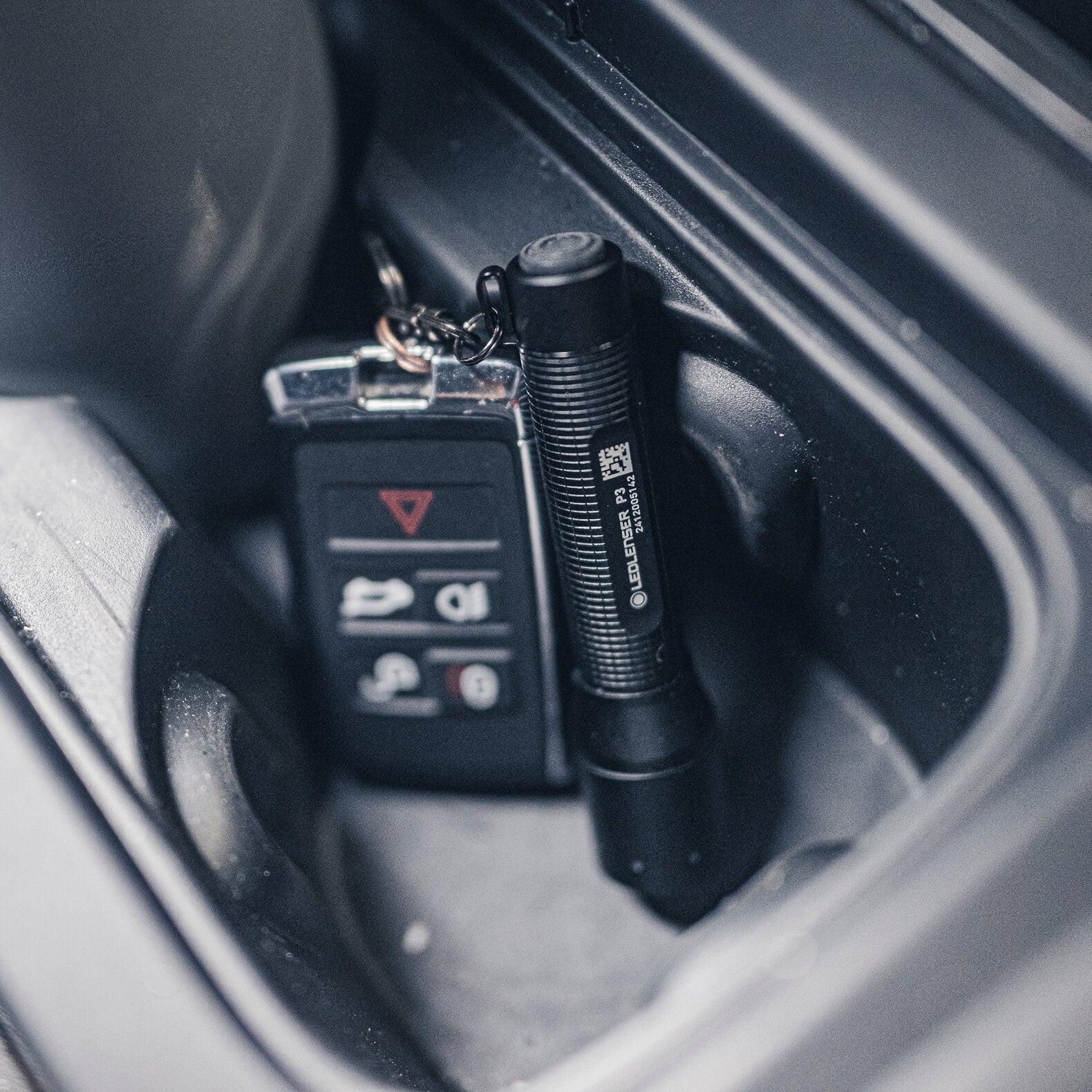 Ein Satz Autoschlüssel und eine kompakte schwarze LEDLENSER Ledlenser Taschenlampe P3 liegen im Getränkehalter eines Autos. Der Schlüsselanhänger hat mehrere Tasten, während die Ledlenser Taschenlampe P3 am Schlüsselbund befestigt ist.