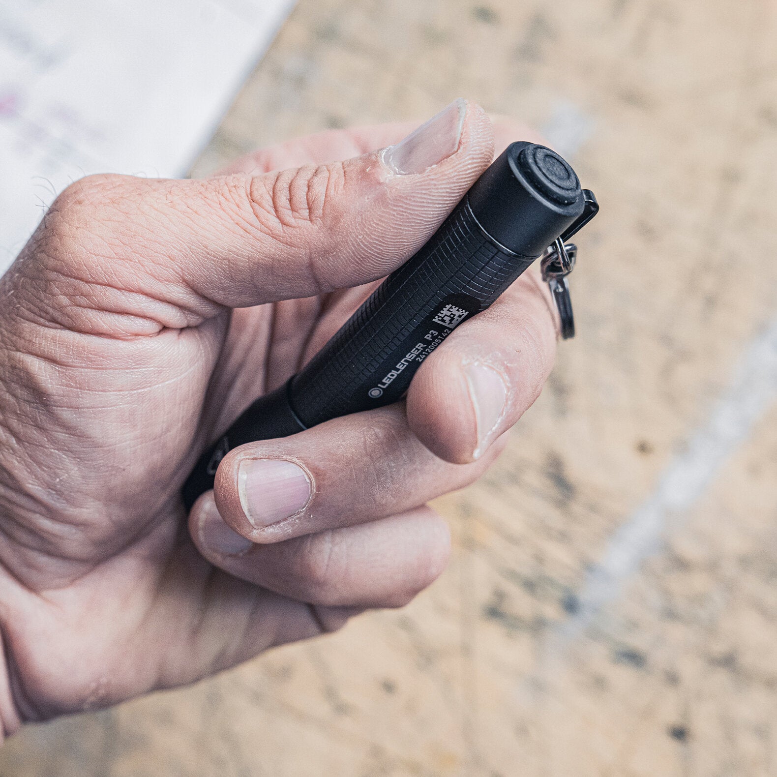 Eine Hand hält die LEDLENSER Ledlenser Taschenlampe P3 mit strukturiertem Griff und Schlüsselbundbefestigung über einer Holzoberfläche - ideal für den täglichen Gebrauch.