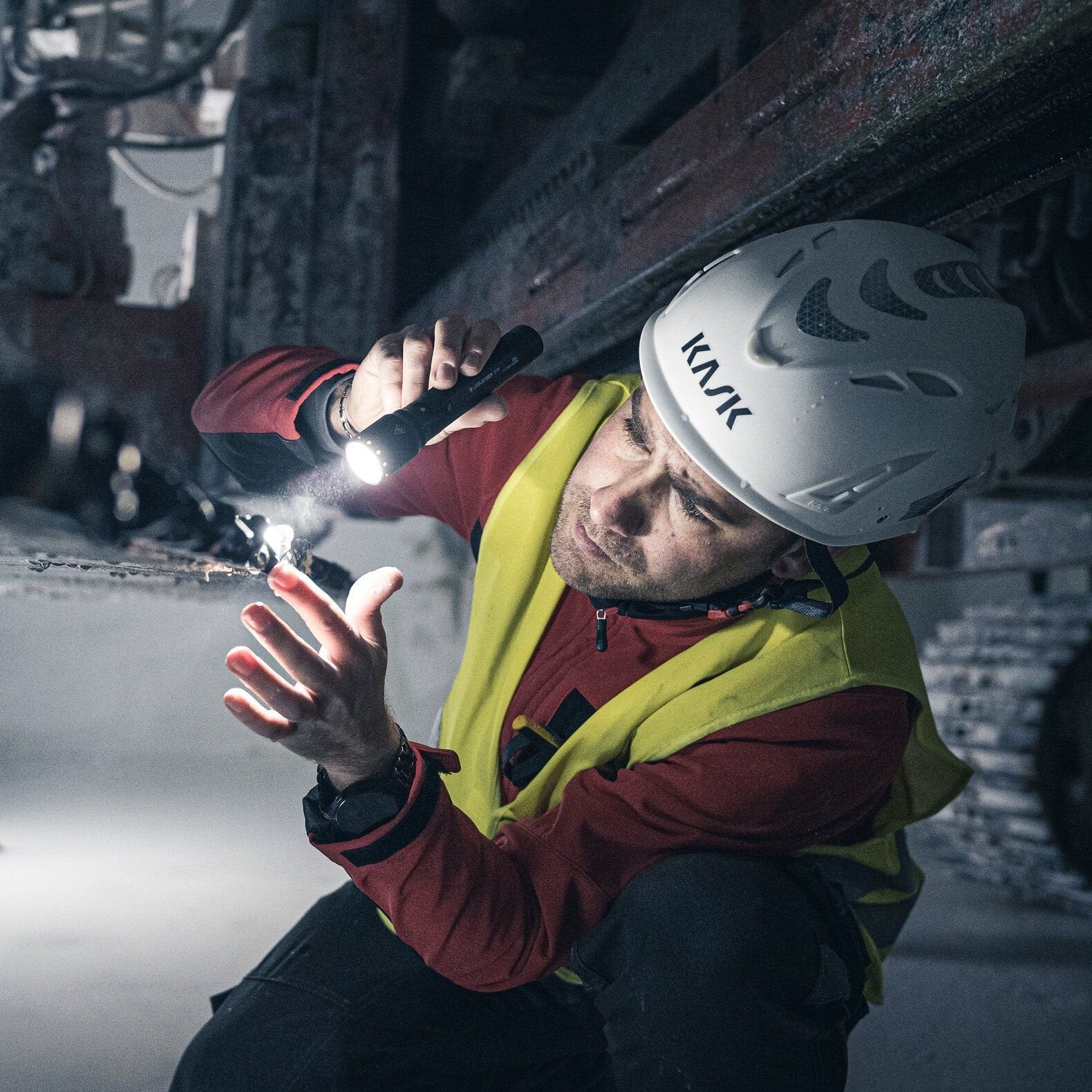 Ein Arbeiter mit gelber Sicherheitsweste und weißem Helm benutzt die LEDLENSER Ledlenser Taschenlampe P7R, um in einer schwach beleuchteten Industrieumgebung Maschinen genau zu inspizieren. Er geht in die Hocke, um eine bessere Sicht zu haben.