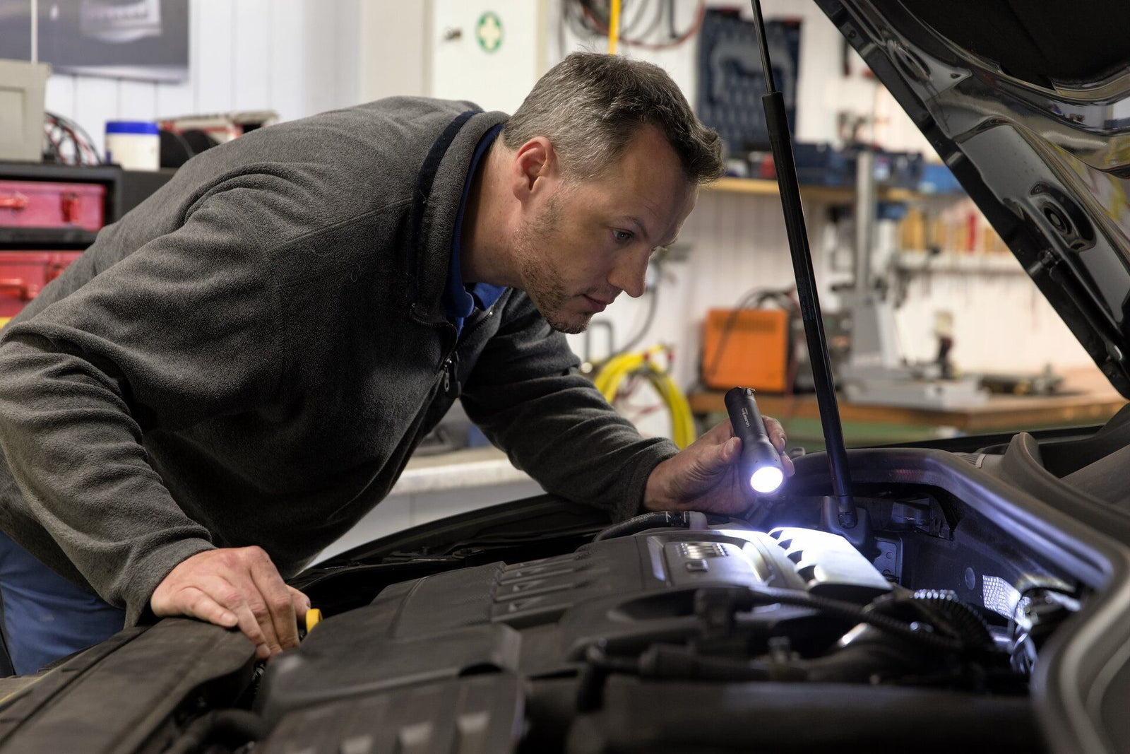 In einer Werkstatt inspiziert ein Mann mit der LEDLENSER Taschenlampe P7 einen Automotor unter der Motorhaube und nutzt das Advanced Focus System, um die Komponenten genau zu untersuchen.
