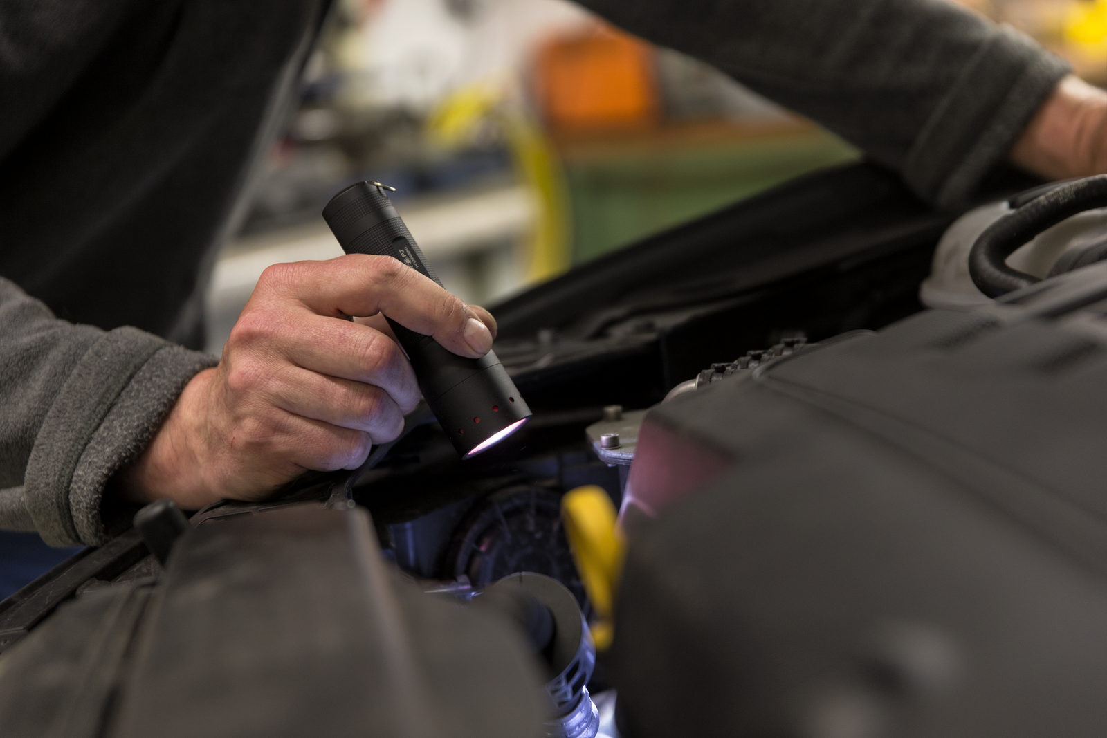 In einer Werkstatt leuchtet eine Person mit der LEDLENSER Ledlenser Taschenlampe P7 mit einer Hand durch das Advanced Focus System in den Motorraum eines Autos, während die andere Hand auf dem Fahrzeug ruht.