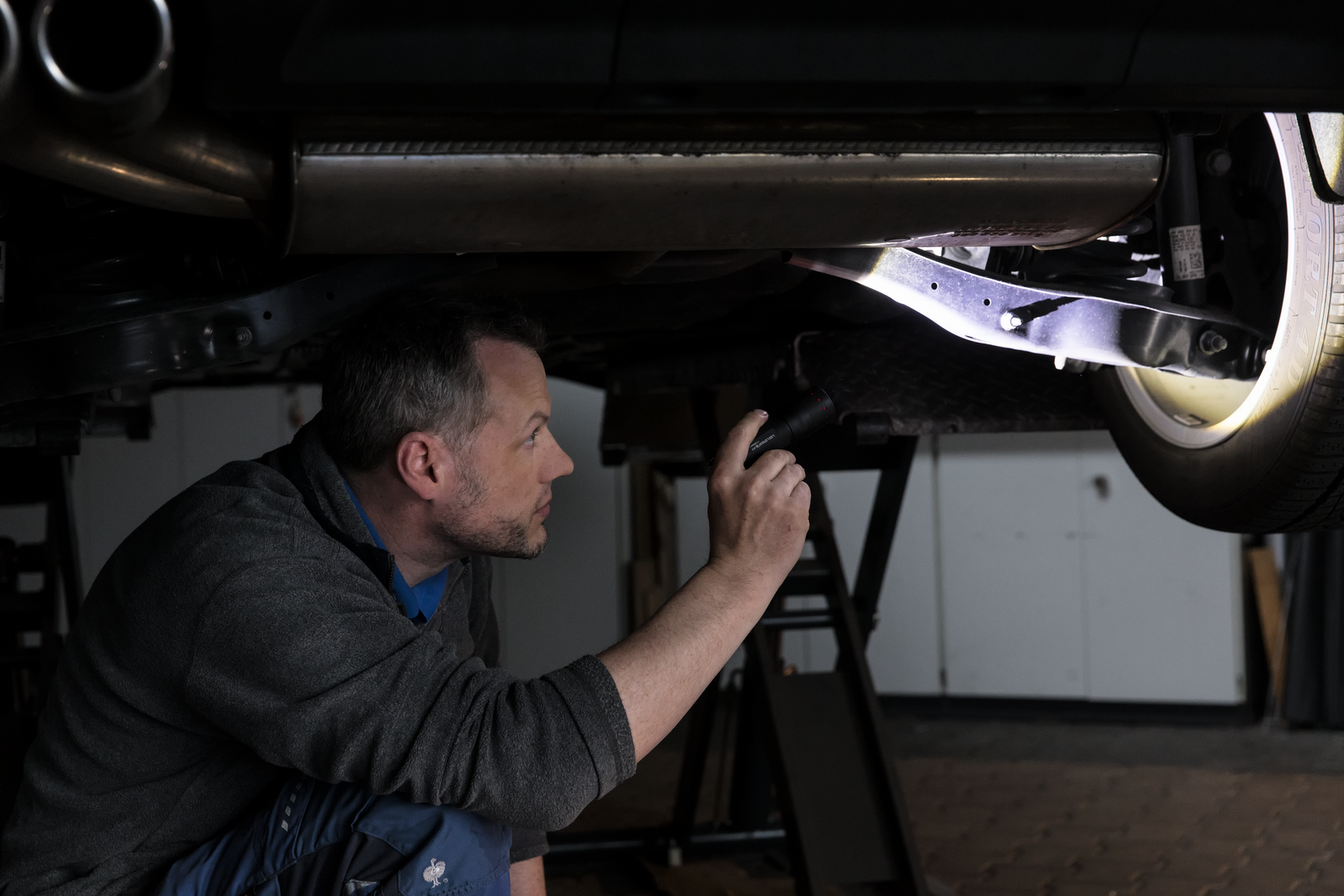 Ein Mann hockt in einer Werkstatt unter einem Auto und inspiziert die Unterseite mit der LEDLENSER Ledlenser Taschenlampe P7.