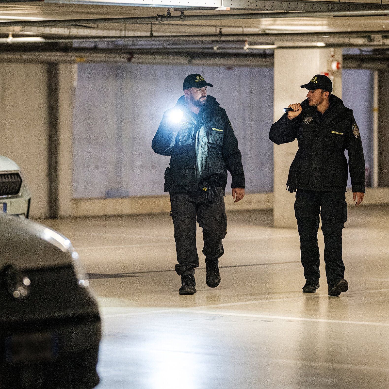 Zwei uniformierte Sicherheitsbeamte gehen durch eine schummrige Tiefgarage und inspizieren den Bereich mit einer LEDLENSER Ledlenser Taschenlampe TT3R. Im Hintergrund sind geparkte Autos zu sehen.