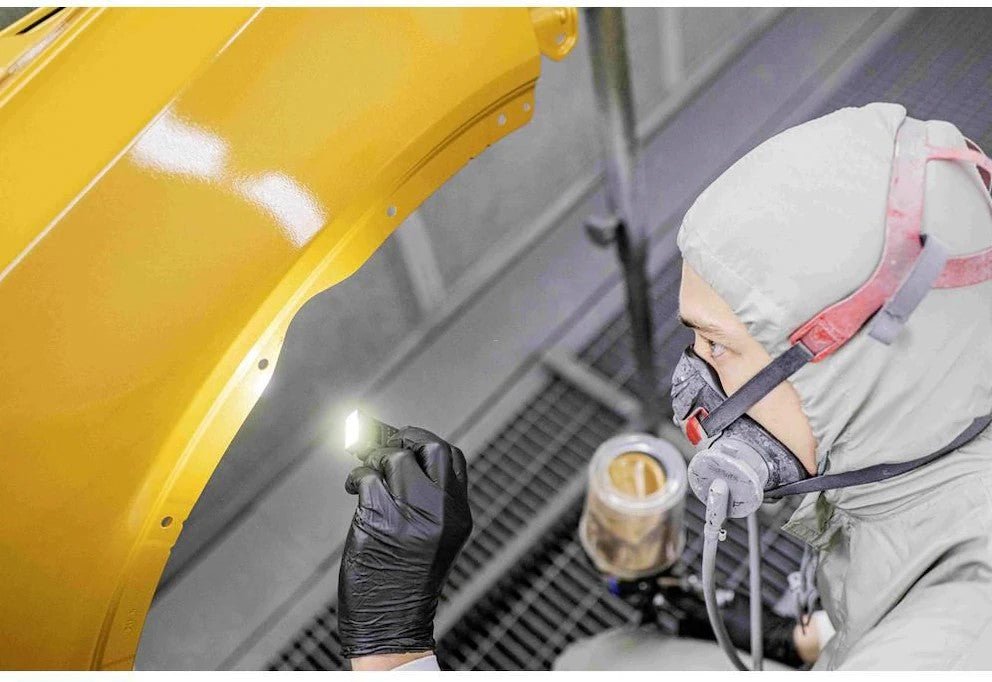 Mit Schutzkleidung und Atemschutzmaske inspiziert eine Person mit der LEDLENSER Ledlenser W1R Work ein gelb lackiertes Autoteil in einer industriellen Umgebung.