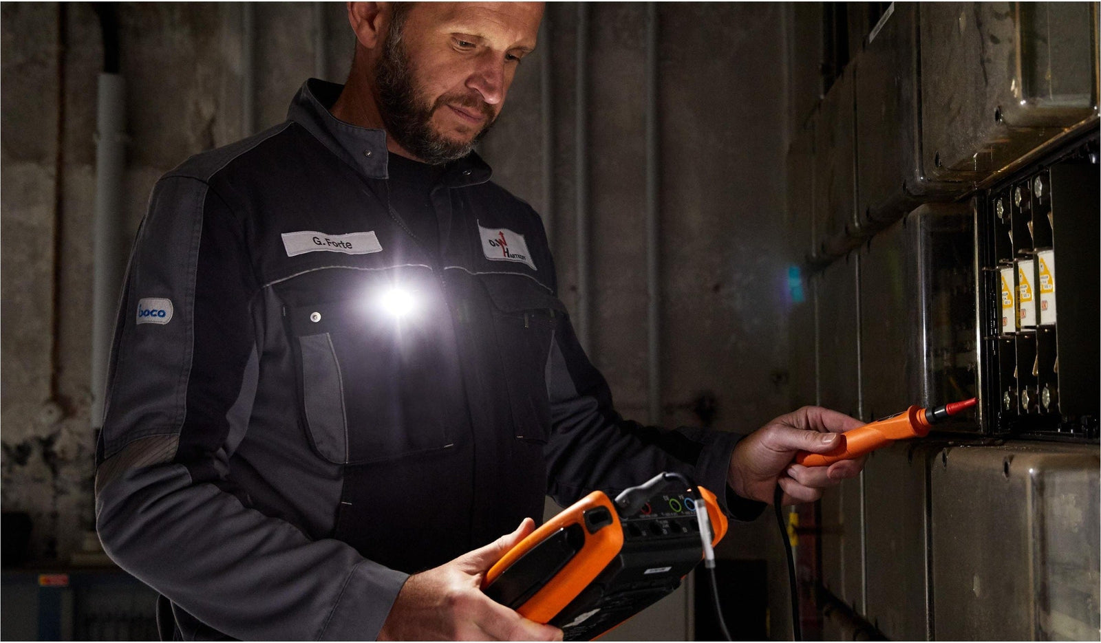 Ein Techniker in dunkler Uniform prüft mit einem orangefarbenen Multimeter eine elektrische Schalttafel. An seiner Brust ist eine LEDLENSER Ledlenser W1R Work befestigt, die seinen Arbeitsbereich in dem schwach beleuchteten Raum hell ausleuchtet.