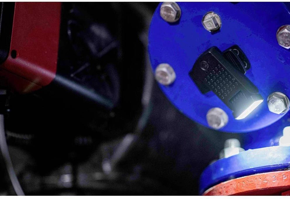 Die kompakte schwarze LEDLENSER Ledlenser W1R Work beleuchtet Schrauben und Maschinen, indem sie auf einen großen blauen Rohrflansch aufgesteckt wird. Sie erhellt einen schummrigen Industrieraum mit konzentriertem, wiederaufladbarem Licht, wo es am meisten gebraucht wird.