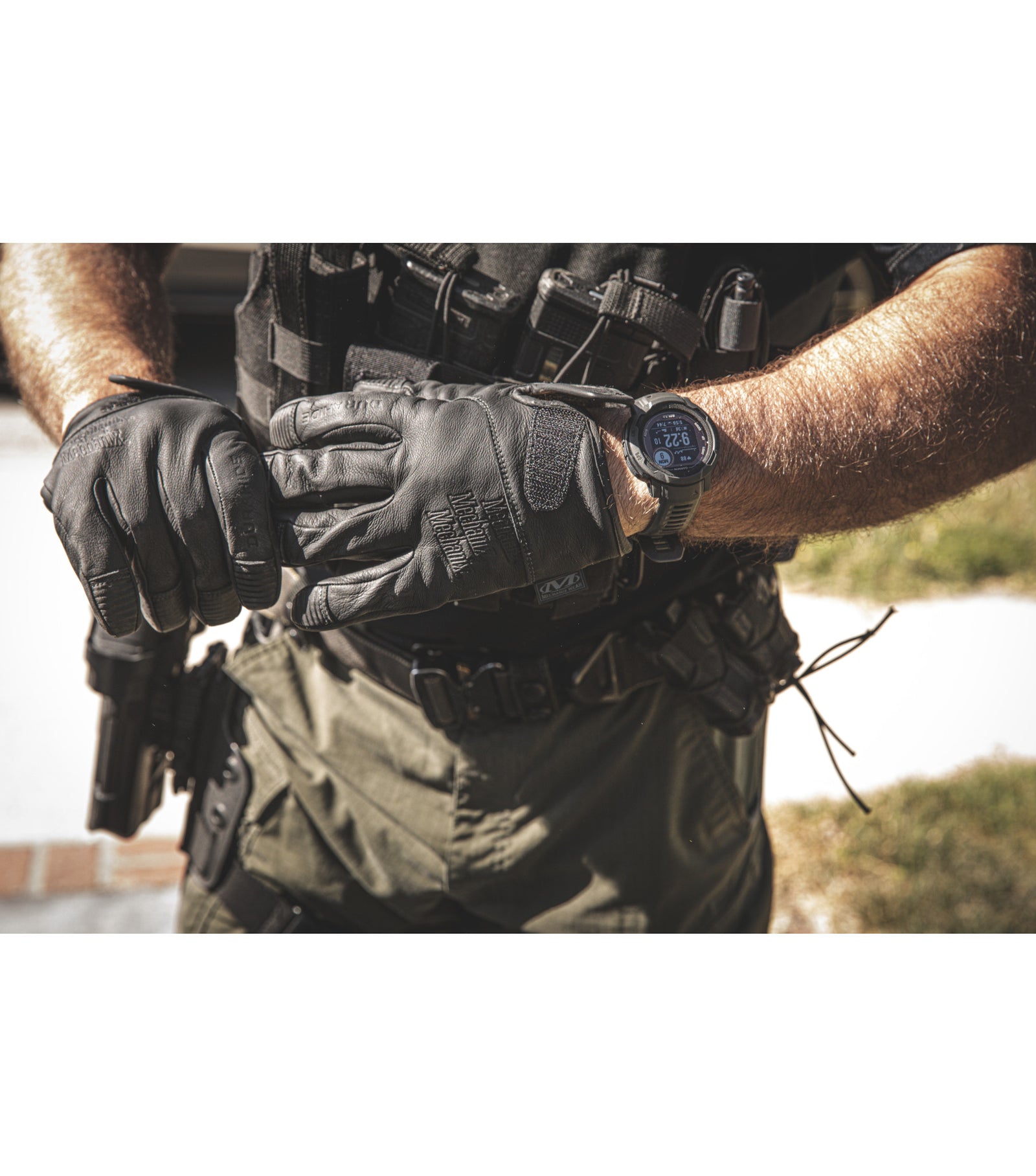 Eine Person, die MECHANIX Mechanix Handschuhe Durahide Needlestick LE - Black Handschuhe, taktische Ausrüstung und einen Durahide Leder Utility Belt trägt, greift eine Schusswaffe im Freien im Sonnenlicht. Nur ihr Oberkörper und ihre Arme sind sichtbar.