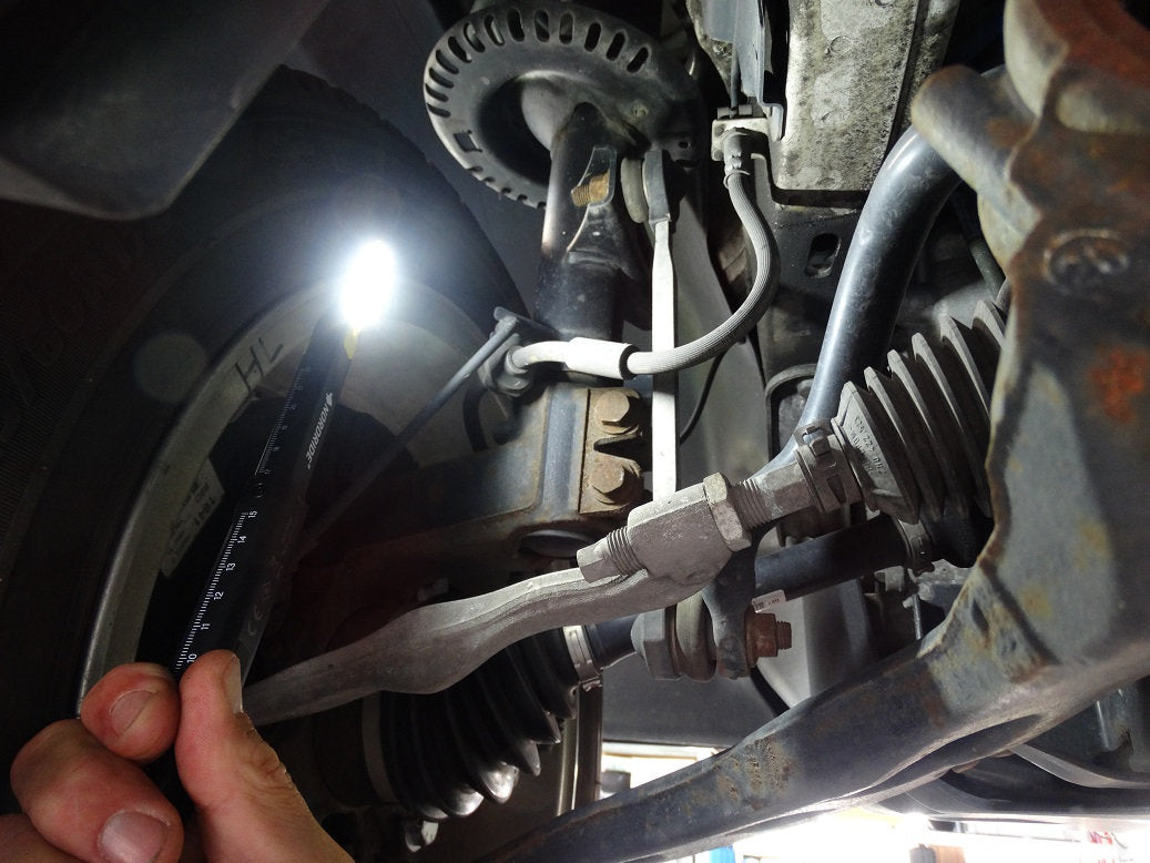 Nahaufnahme der Aufhängung und Lenkung eines Fahrzeugs, wobei eine Person die NORDRIDE Nordride Arbeitslampe Pen Light Flex und ein Lineal in der Nähe der Radnabe verwendet, um die Komponenten zu prüfen.