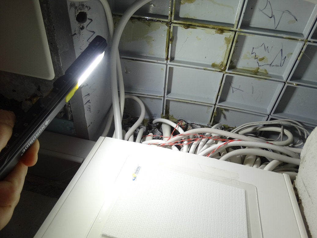Eine Hand, die eine NORDRIDE Nordride Arbeitslampe Pen Light Flex hält, beleuchtet hell die weißen und roten elektrischen Drähte über einer Deckenplatte in einem schwach beleuchteten Raum mit markierten Deckenplatten.