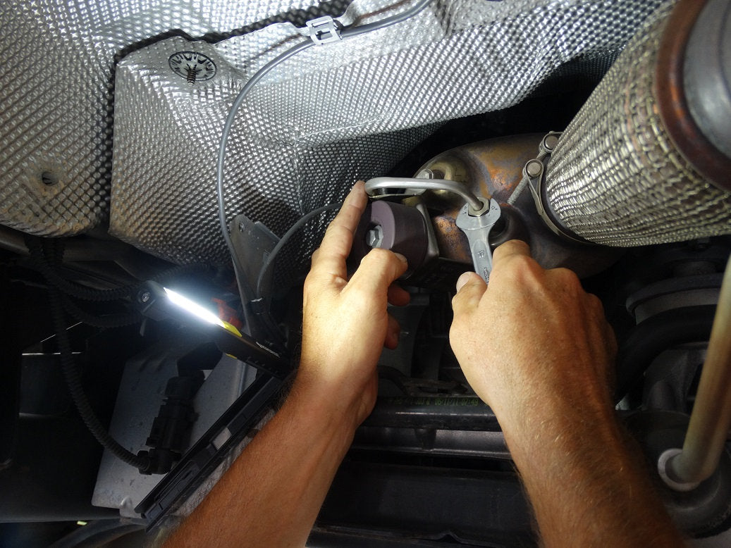 Eine Person arbeitet mit einem Schraubenschlüssel an einem Metallteil unter einem Fahrzeug, das von der NORDRIDE Nordride Arbeitslampe Pen Light Flex mit Magnetclip beleuchtet wird, während verschiedene Drähte und Hitzeschilde sichtbar sind.