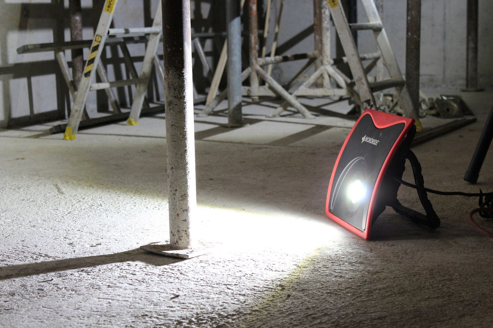 Der NORDRIDE Nordride Arbeitsstrahler Power Light 60W mit rotem Rahmen leuchtet einen Betonboden mit 5000 Lumen hell aus und wirft Licht auf umliegende Leitern und Metallgerüste.
