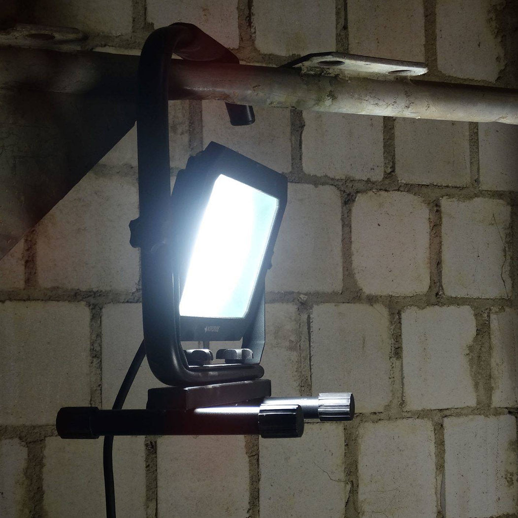 Ein NORDRIDE Nordride Baustrahler Sector 30W IP65 ist an ein Metallrohr geklemmt und beleuchtet hell eine weiße Betonblockwand in einer Industriehalle. Die robuste Schutzart IP65 sorgt für zuverlässige Beleuchtung unter rauen Bedingungen.