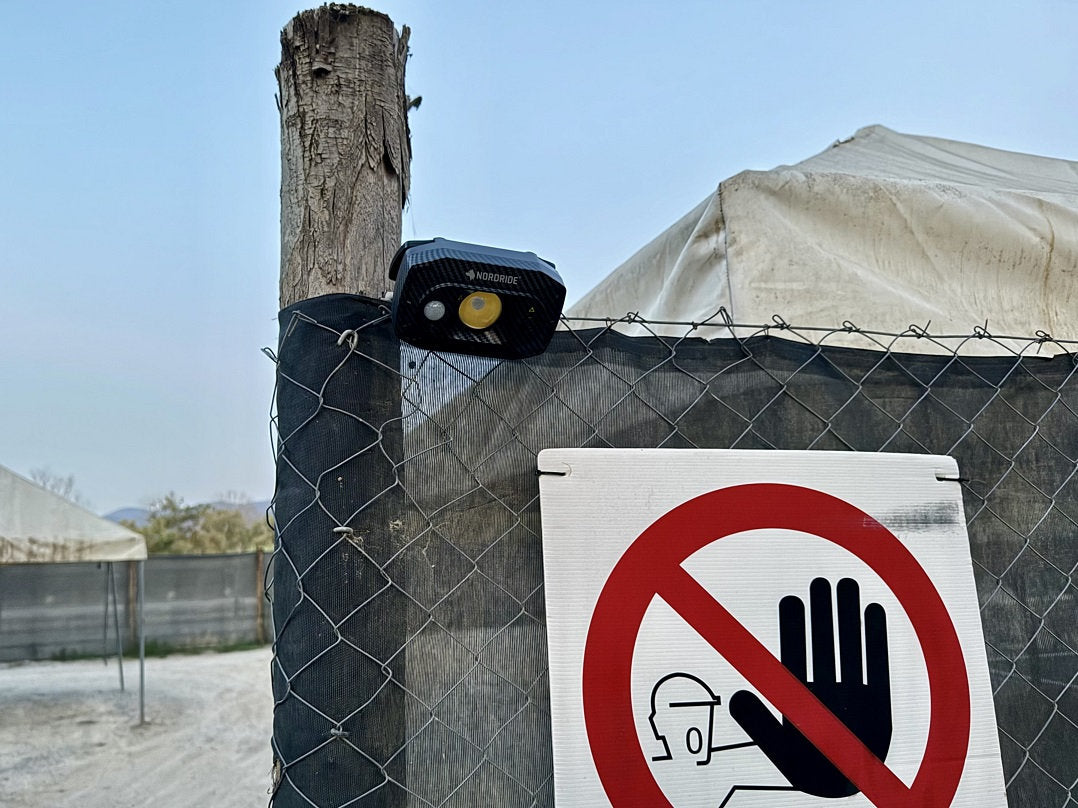 Ein NORDRIDE Nordride Flutlichtstrahler Guard mit IP67-Schutz ist an einem Holzpfosten über einem Maschendrahtzaun montiert. Darunter weist ein Schild auf Zutrittsbeschränkung hin, im Hintergrund sind Zelte sichtbar.