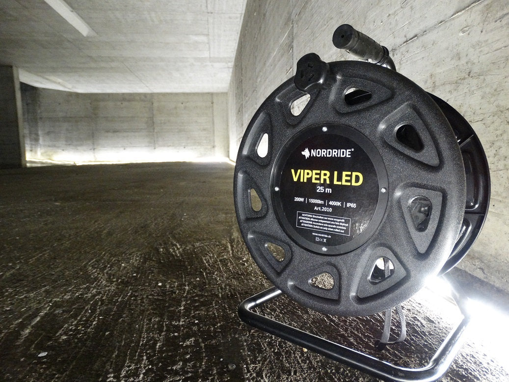 Eine NORDRIDE Lichtband Viper LED 25m Kabeltrommel, perfekt für die Baustellenbeleuchtung, wird auf einem Betonboden in einem großen, leeren, schwach beleuchteten Raum mit grauen Wänden platziert. Seine hellen LED-Leuchten erhellen den Raum.