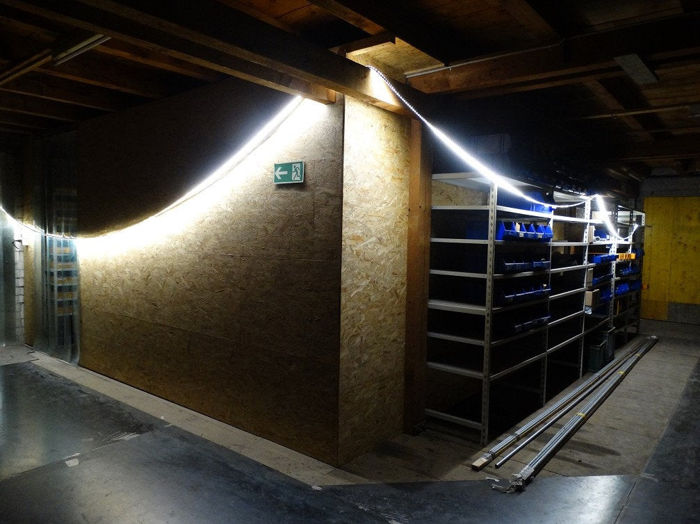 Ein schummrig beleuchteter Lagerraum mit Holzwänden, Metallregalen und blauen Behältern. Ein NORDRIDE Lichtband Viper LED 25m leuchtet an der Decke. Ein Notausgangsschild befindet sich an einer Wand und Metallrohre sind auf dem Boden gestapelt.