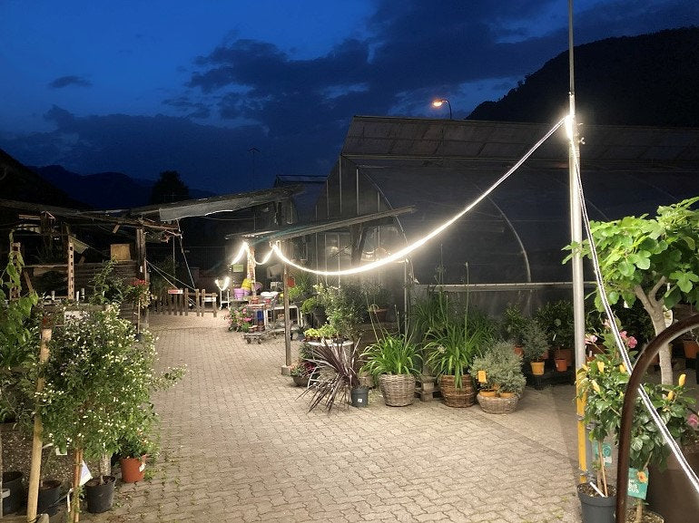 In der Abenddämmerung erstrahlt das Gartencenter im Freien mit Lichterketten und dem NORDRIDE Lichtband Viper LED 25m, das die Topfpflanzen entlang des Weges beleuchtet. Gewächshäuser und Bergansichten werden unter Baustellenbeleuchtung gegen den dunkler werdenden Himmel hervorgehoben.