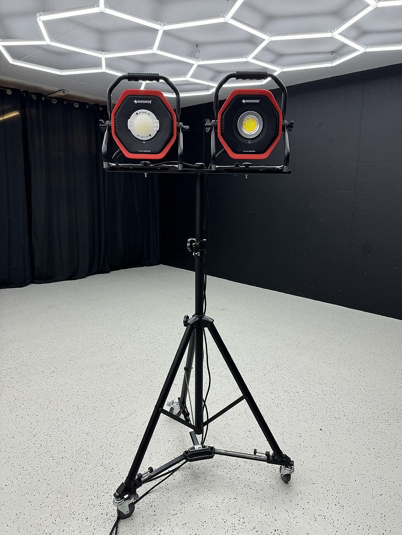 Zwei quadratische LED-Arbeitsleuchten sind an der NORDRIDE Nordride Stativ in einem modernen Raum mit schwarzen Wänden, schwarzen Vorhängen, einem weiß gesprenkelten Boden und geometrischen Deckenleuchten angebracht.