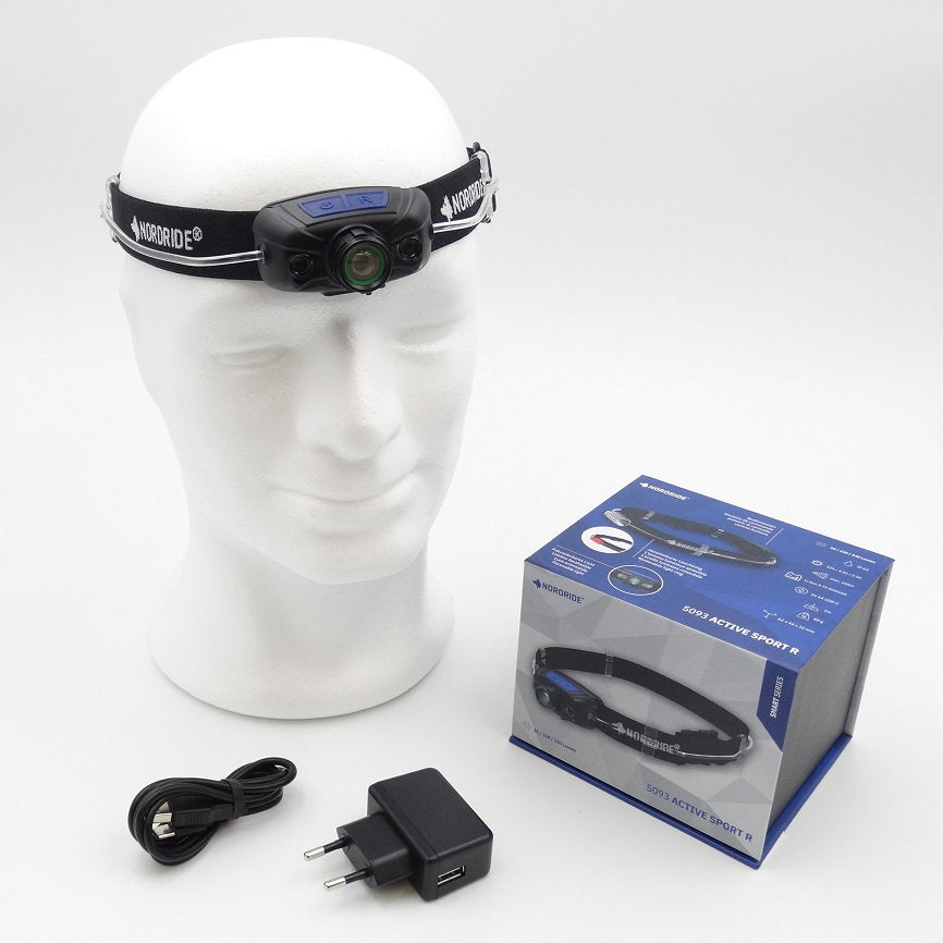 Eine weiße Schaufensterpuppe trägt eine NORDRIDE Stirnlampe Active Sport R. Daneben liegen ein USB-Kabel, ein Ladestecker und die blaue Verkaufsverpackung der wiederaufladbaren Stirnlampe mit Produktbildern und Details.