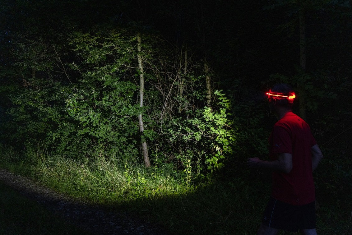 Eine Person, die die NORDRIDE Strinlampe Active Sport R mit Rotlicht trägt, geht nachts einen dunklen Waldweg entlang und beleuchtet mit dem Licht der Lampe das dichte grüne Laub vor sich.