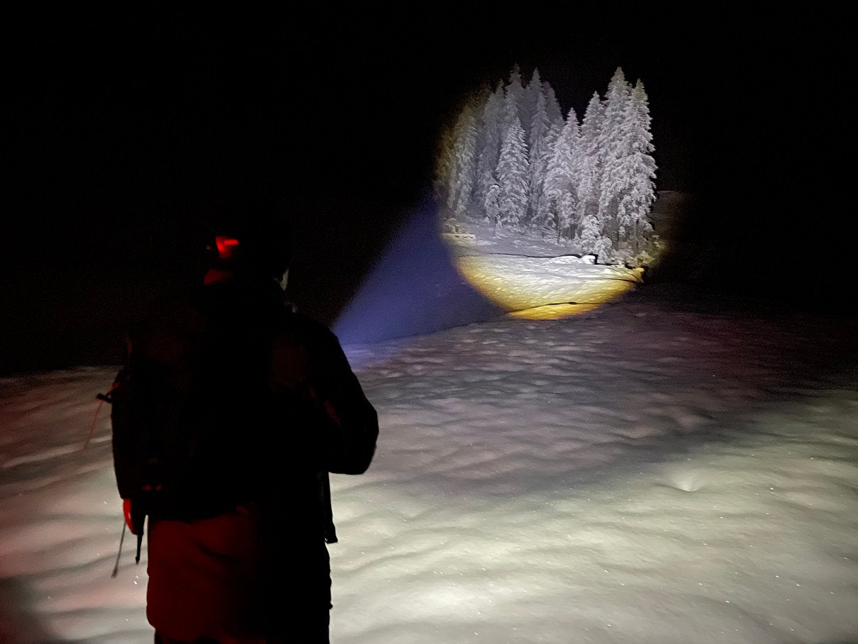 Eine Person in einer verschneiten Nachtlandschaft benutzt die NORDRIDE Nordride Taschenlampe Spot Long Ranger R, deren kräftiger Lichtstrahl mit 1000m Reichweite weit entfernte, schneebedeckte Bäume gegen die Dunkelheit beleuchtet.