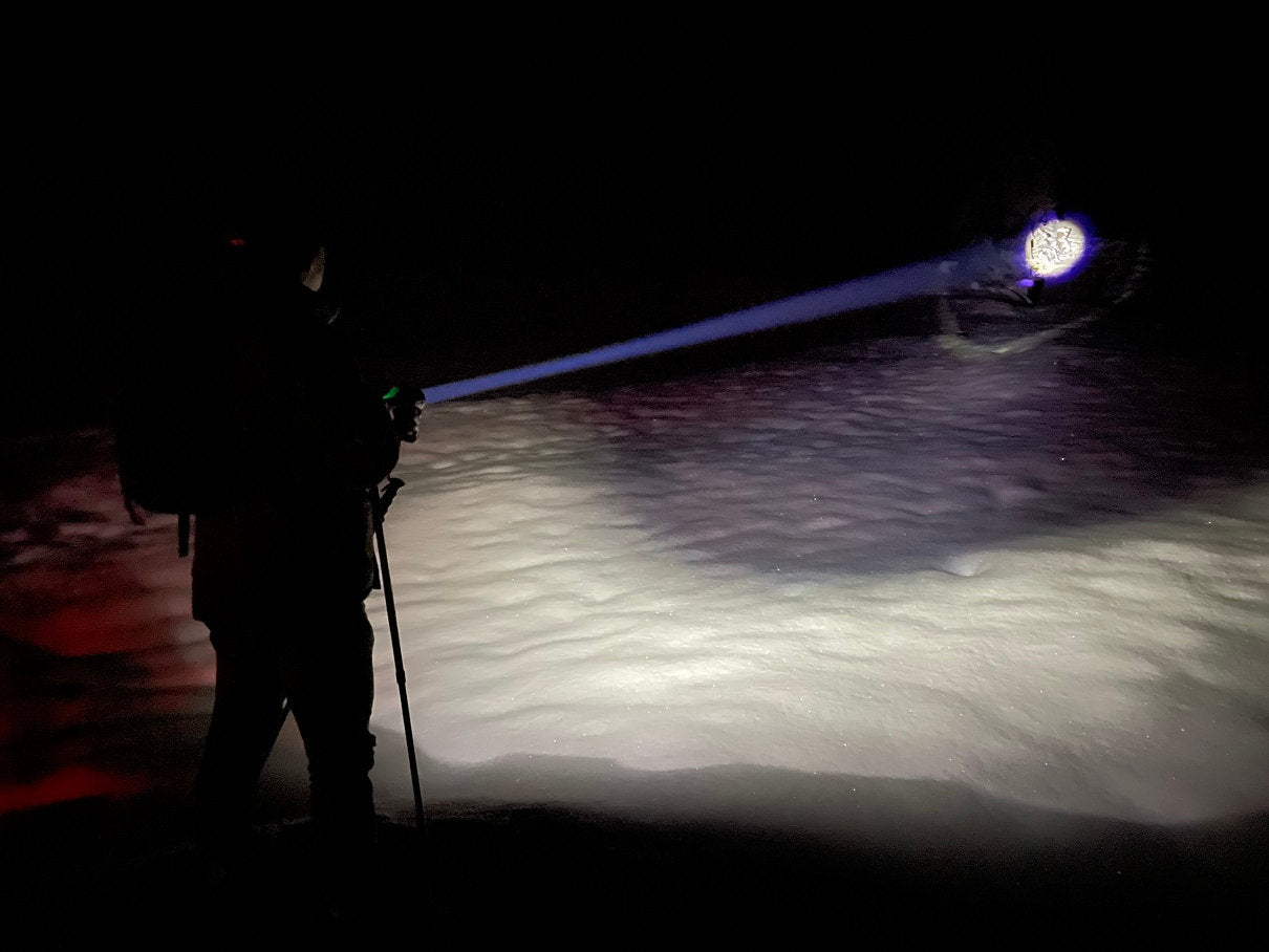 Eine Person mit Gehstock benutzt die NORDRIDE Nordride Taschenlampe Spot Long Ranger R und wirft in der Dunkelheit einen hellen Lichtstrahl über den verschneiten Boden, während die Umgebung meist im Schatten bleibt.