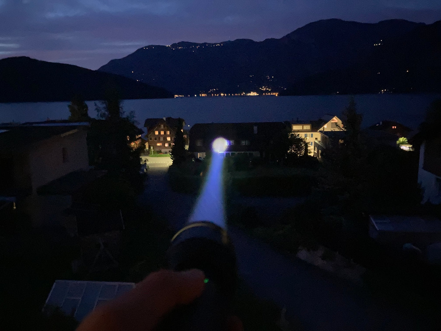 Eine Hand hält eine NORDRIDE Nordride Taschenlampe Spot Long Ranger R, deren kräftiger Lichtstrahl Häuser und einen nächtlichen See erhellt, Berge im Hintergrund und ferne Lichter auf dem Wasser, die die Reichweite von 1000 Metern verdeutlichen.