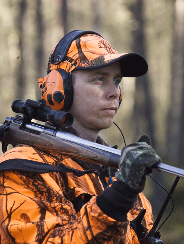 Bekleidet mit orangefarbenem Tarnanzug, Mütze und 3M Peltor Gehörschutz SportTac steht ein Jäger im Wald, mit dem Gewehr auf der Schulter und konzentriertem Blick - bereit für die Jagd.