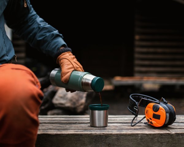 Eine Person in einem orangefarbenen Handschuh gießt ein heißes Getränk aus einer grünen Thermoskanne in einen Metallbecher auf Holz, neben dem Becher stehen ein orangefarbenes Kofferradio und ein 3M Peltor Gehörschutz SportTac Gehörschutz.