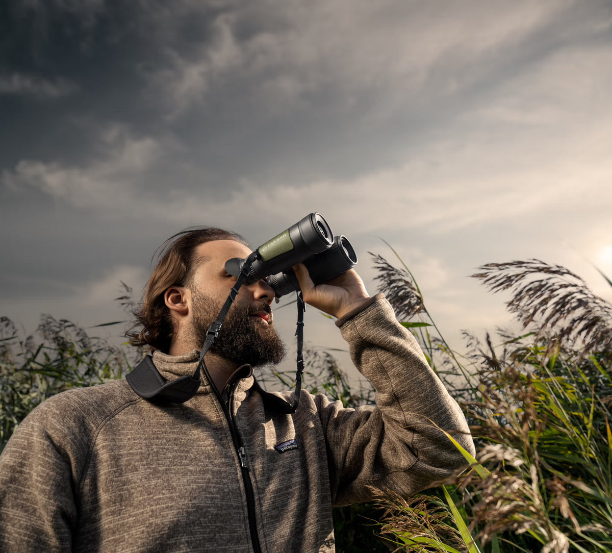 Ein bärtiger Mann in einer grauen Jacke benutzt das PULSAR Pulsar WLO Wärmebildfernglas Ventex XP35, um die Natur zu beobachten. Er steht im hohen Gras unter einem wolkenverhangenen Himmel, was an Vogel- oder Wildtierbeobachtung denken lässt.