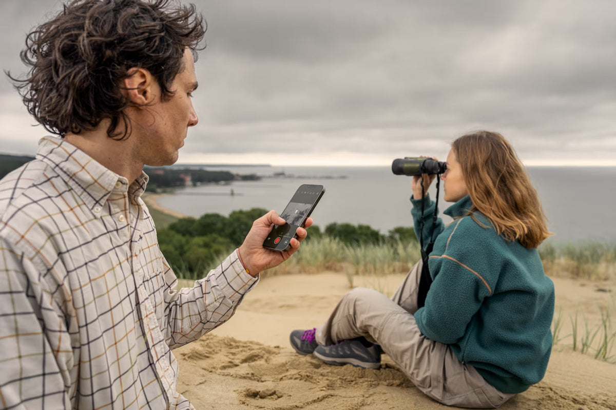 Ein Mann mit einem Smartphone steht auf sandigem Boden und beobachtet eine Frau in der Nähe, die mit dem PULSAR WLO Wärmebildfernglas Ventex XP35 das Meer und die Küstenlinie unter einem bewölkten Himmel beobachtet.