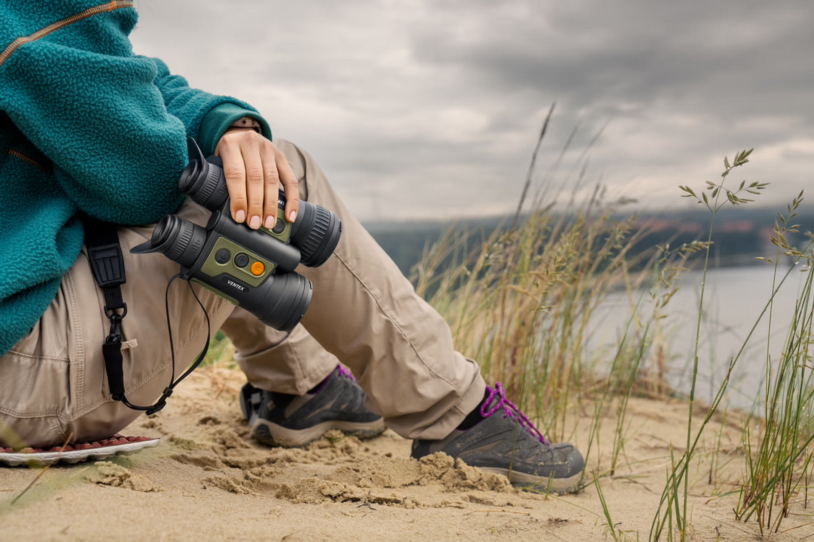 Eine Person, die nur durch ihre Hand, ihre Beine und einen Teil ihrer grünen Jacke erkennbar ist, sitzt auf sandigem Boden in der Nähe von hohem Gras und hält ein PULSAR WLO Wärmebildfernglas Ventex XT50. Der Hintergrund zeigt einen bedeckten Himmel und Wasser.