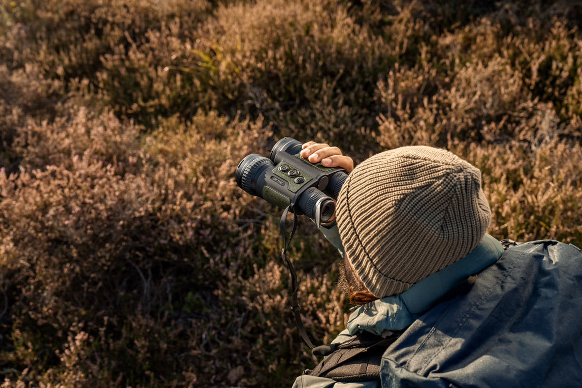 Eine Person mit beiger Strickmütze und Outdoor-Jacke ist von hinten zu sehen, die ein PULSAR WLO Wärmebildfernglas Ventex XT50 in der Hand hält und auf eine grasbewachsene, sonnenbeschienene Landschaft blickt.