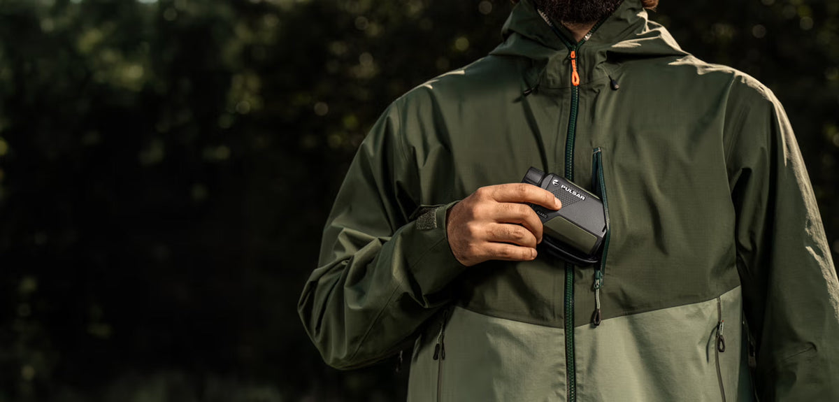 Eine Person in grüner Outdoor-Jacke hält sich ein schwarzes PULSAR Pulsar WLO Wärmebildgerät Alaris XG30 vor die Brust. Der Hintergrund ist mit dunklem Laub unscharf.