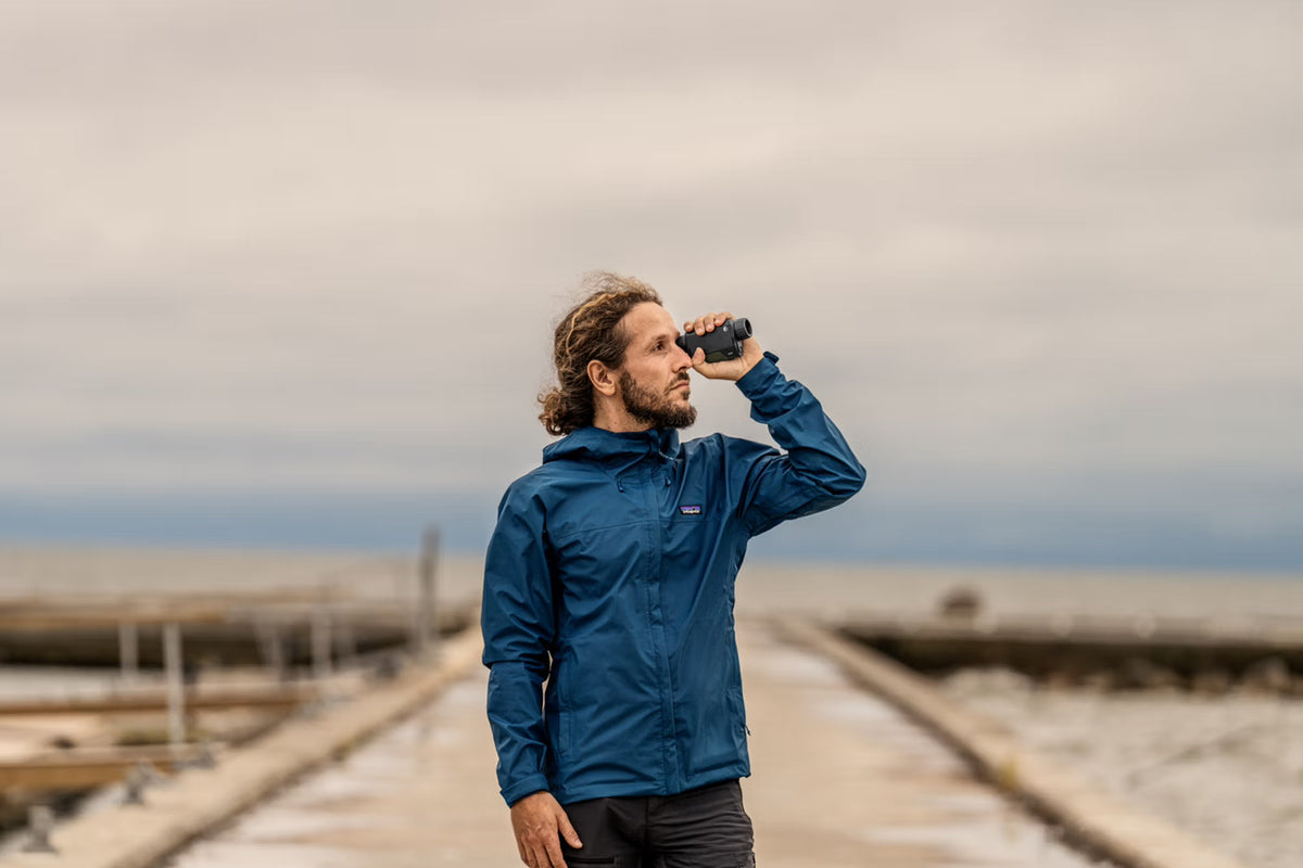 Ein Mann in einer blauen Jacke steht auf einem Steg am Wasser und schaut durch ein PULSAR Pulsar WLO Wärmebildgerät Alaris XQ30 auf den Horizont unter einem bewölkten Himmel.