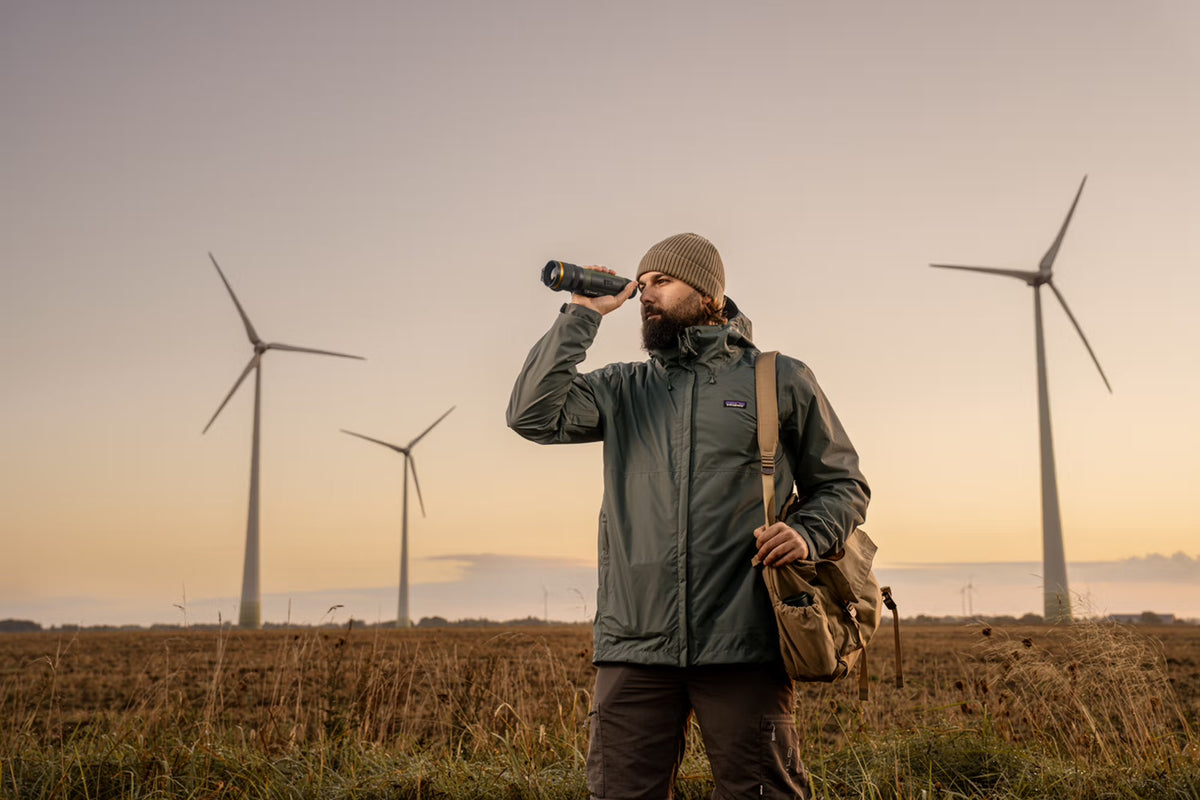 Ein Mann in Outdoor-Kleidung mit Mütze und Umhängetasche steht bei Sonnenuntergang auf einem Feld und schaut durch sein PULSAR Pulsar WLO Wärmebildgerät Lumion XL50, während sich im Hintergrund mehrere Windräder erheben.