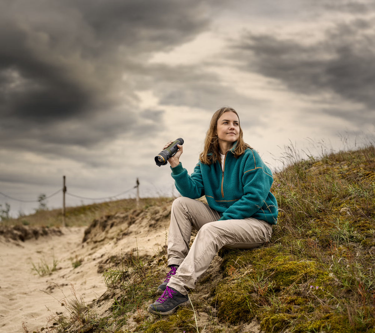 Eine Frau in Outdoor-Ausrüstung sitzt auf einer grasbewachsenen Anhöhe in der Nähe eines Sandweges, hält ihr PULSAR Pulsar WLO Wärmebildgerät Lumion XL50 in der Hand und schaut in die Ferne unter einem bewölkten Himmel.