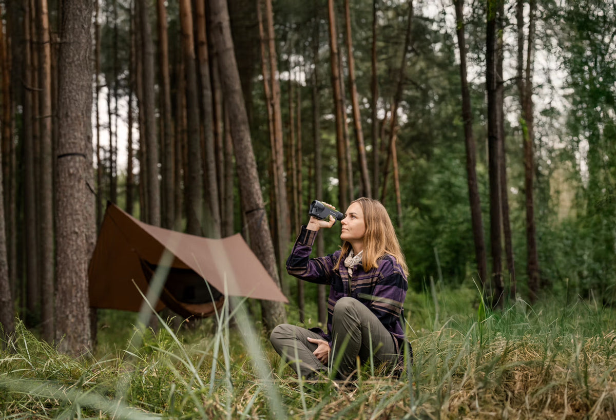 Eine Frau in Outdoor-Kleidung genießt ein heißes Getränk auf der Wiese mit ihrem PULSAR Pulsar WLO Wärmebildgerät Orni XG35 neben ihrem Zelt, perfekt für Natur- und Wildtierbeobachtung im ruhigen Wald unter hohen Bäumen.