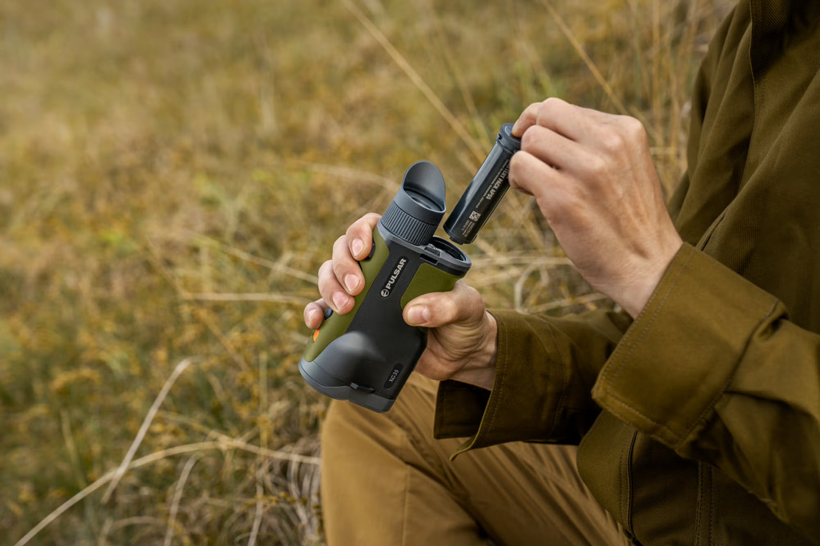 Eine Person in brauner Outdoor-Ausrüstung setzt eine Batterie in ein grün-schwarzes PULSAR WLO Wärmebildgerät Orni XG35 ein, während sie im Gras sitzt und sich auf die Natur- und Tierbeobachtung in einer friedlichen Umgebung vorbereitet.