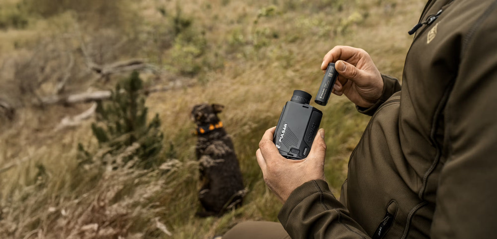 Eine Person in Outdoor-Kleidung hält ein PULSAR Wärmebildgerät Axion XQ19 Compact und eine Batterie in der Hand und bereitet das Einsetzen vor, während ein Hund mit einem orangefarbenen Halsband in der Nähe im hohen Gras sitzt.