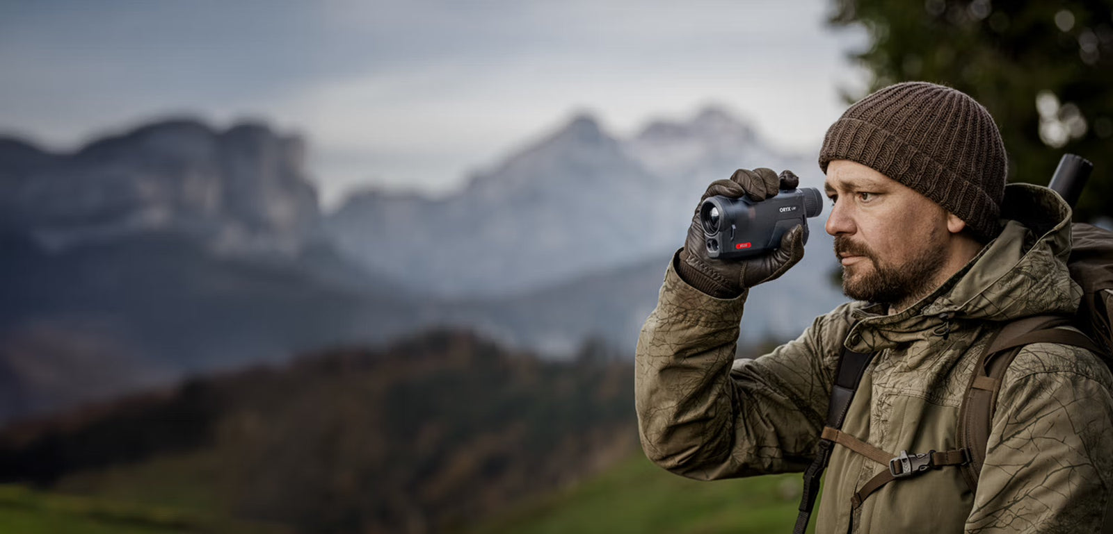 Ein Mann in Outdoor-Kleidung und Strickmütze benutzt das PULSAR Pulsar Wärmebildgerät Oryx LRF XG35, während er in einer bergigen Landschaft steht - ideal für die Jagd - mit verschwommenen Gipfeln als dramatischer Kulisse.