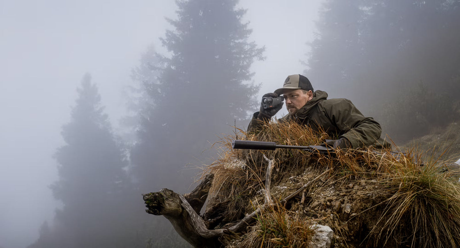 Ein Jäger in grüner Jacke hockt hinter felsigem Gelände in einem nebligen Wald und benutzt das PULSAR Pulsar Wärmebildgerät Oryx LRF XG35, während sein entschärftes Gewehr in der Nähe ruht - bereit für jede Jagd.