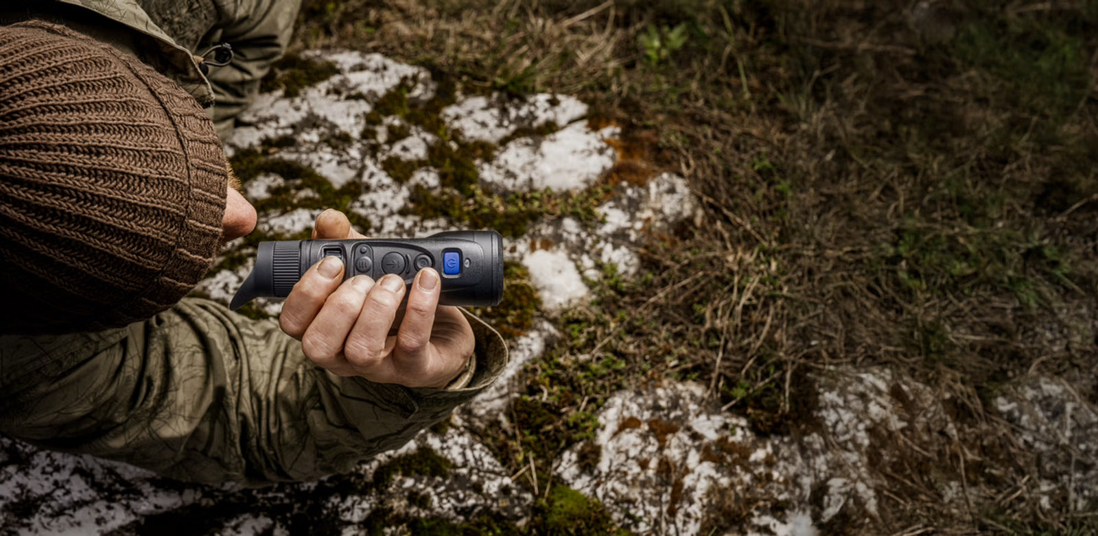 Eine Person in Outdoor-Kleidung hält das kompakte Pulsar Wärmebildgerät Oryx LRF XG35 von PULSAR mit blauen Tasten und integriertem Laser-Entfernungsmesser und richtet es auf den verschneiten, moosigen Waldboden - ideal für die Jagd.