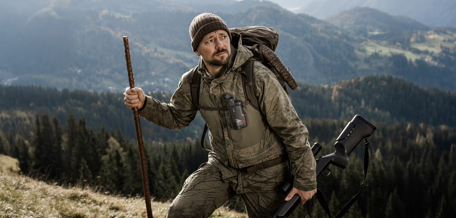 Ein Mann in Tarnkleidung und mit Strickmütze wandert durch malerische bewaldete Hügel und trägt ein Gewehr, einen Rucksack und das PULSAR Pulsar Wärmebildgerät Oryx LRF XG35.