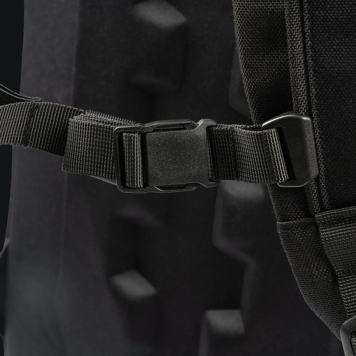 Nahaufnahme einer schwarzen Kunststoffschnalle, die an einem schwarzen Gurt befestigt ist, der an der gepolsterten Stoffoberfläche des STOIRM 25L Tactical Pack GEN2 angebracht ist.