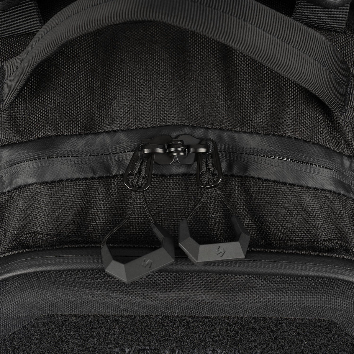 Nahaufnahme des STOIRM 25L Tactical Pack GEN2 in Schwarz, mit zwei Reißverschlüssen mit geometrischen Gummigriffen, teilweise geöffnet in der Nähe eines stabilen Griffs und detaillierten Nähten.