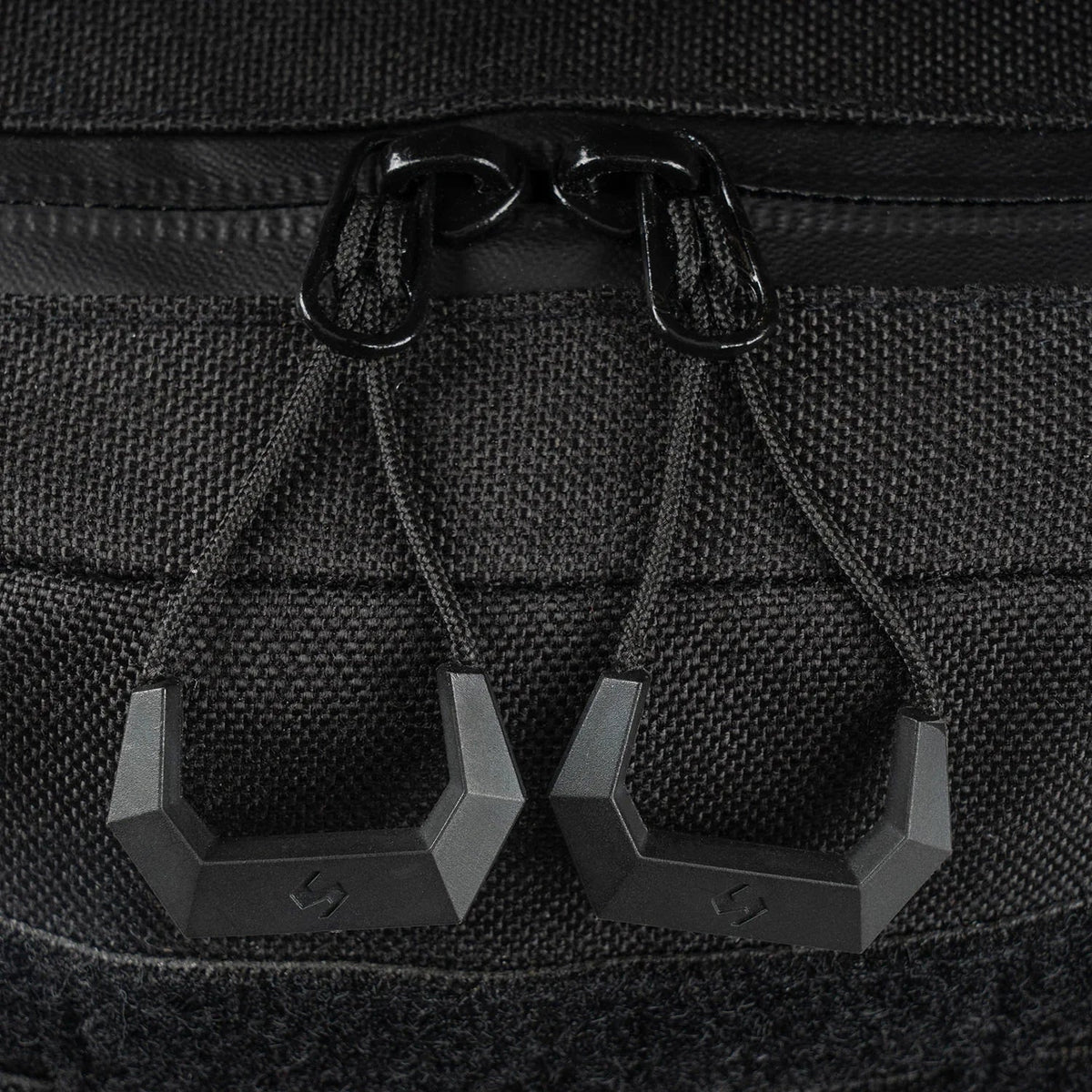Nahaufnahme von zwei schwarzen, sechseckigen Reißverschlüssen mit Kordeln und einem geometrischen Logo auf dem strukturierten Stoff des STOIRM 40L Tactical Pack GEN2 von STOIRM.