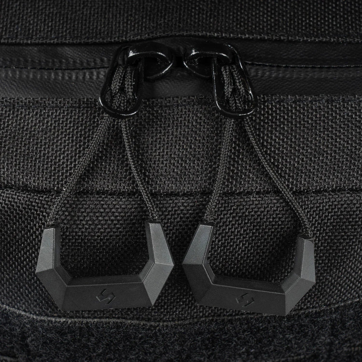 Nahaufnahme des STOIRM 40L Tactical Pack GEN2 von STOIRM. Hervorzuheben sind zwei mattschwarze, geometrische Reißverschlüsse an einem doppelten Schieberegler mit Paracord, umgeben von MOLLE-Gurtband und strapazierfähigem 1000D-Nylongewebe.