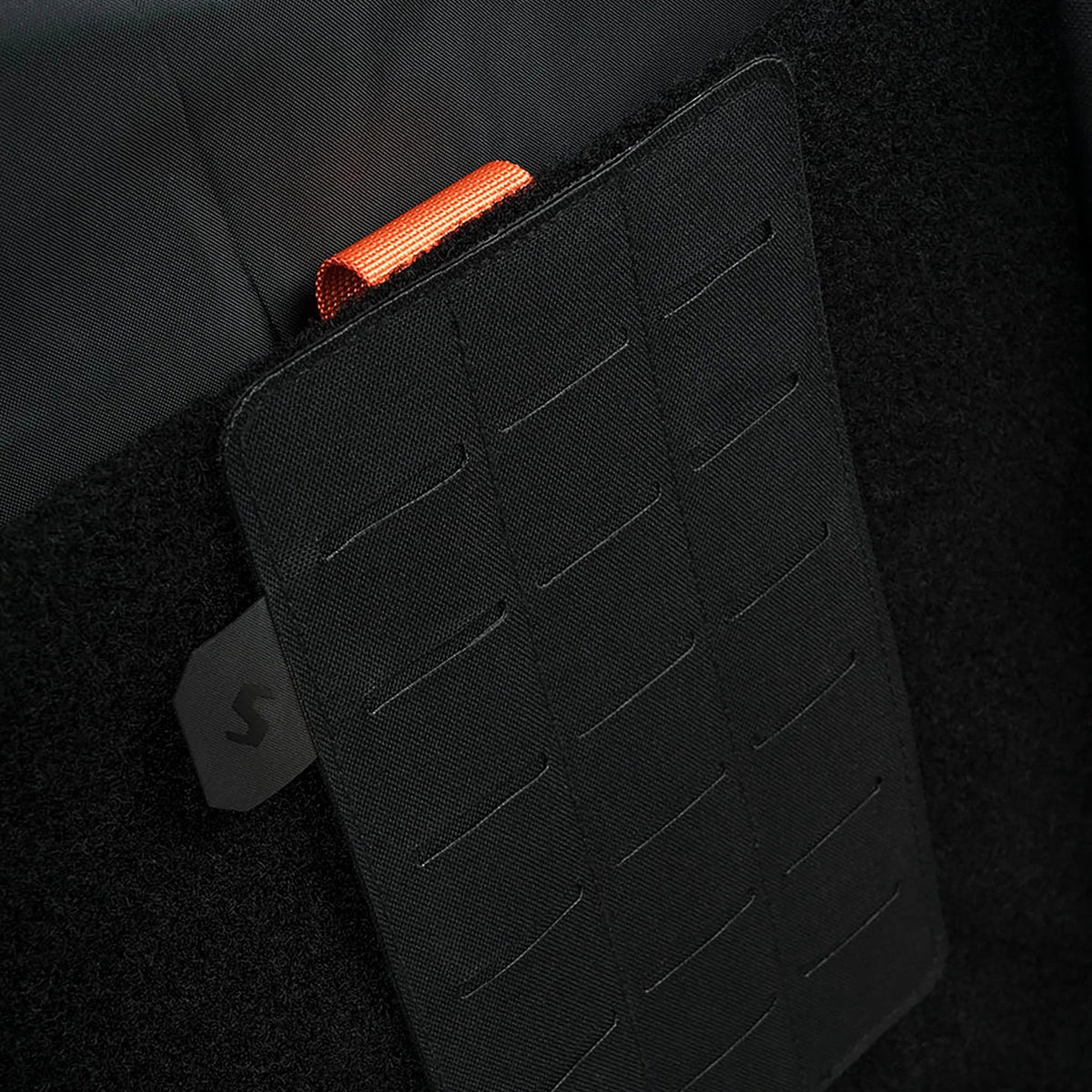 Eine Nahaufnahme des STOIRM Large Molle Panel zeigt den schwarzen, strukturierten Stoff mit horizontalen Schlitzen, eine orangefarbene Zuglasche auf der Oberseite und ein schwarzes STOIRM Eco-System Logo-Tag auf der linken Seite.